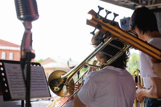 Rear view of young brass section performing on outdoor stage, with trombone player in focus, shining instrument details, bass guitar headstock, and fellow musician adding lively concert energy in