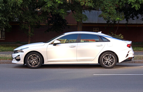 Vinnytsia, Ukraine; April 18, 2026. White KIA K5 2023 at the parking. Sleek, midsize sedan KIA K5 in city street. Korean business car KIA K5 parked on asphalt beside green trees in urban street settin