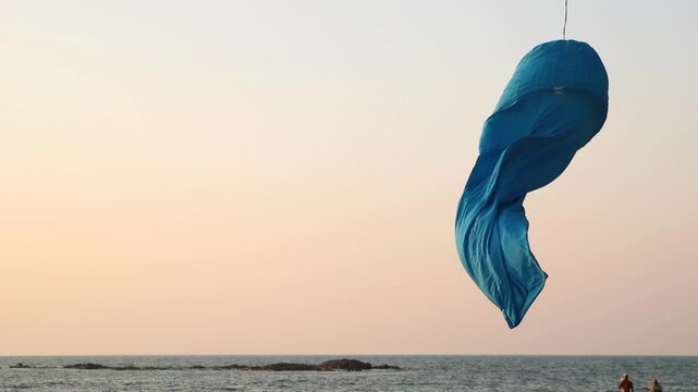 A blue fabric windsock flutters over the sea at sunset over the coastal horizon