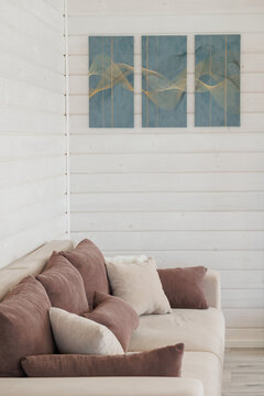 Cozy neutral living room corner featuring beige upholstered sofa layered with multiple soft brown and cream cushions, positioned against white horizontal shiplap wood paneling; three-panel abstract