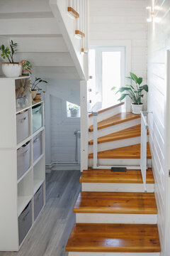 Interior hallway with polished wooden spiral staircase, tall landing window, white paneled walls, built-in shelving with fabric bins, potted plants and soft natural light in modern home