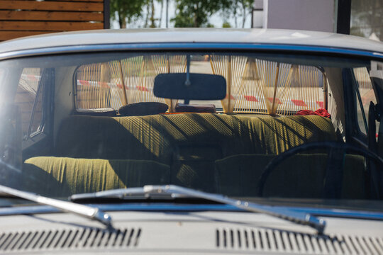 Rear cabin of vintage car viewed through windshield showing olive green fabric bench seat, bamboo string sunshade, hanging mirror, small cushions, red cloth and soft daylight reflections
