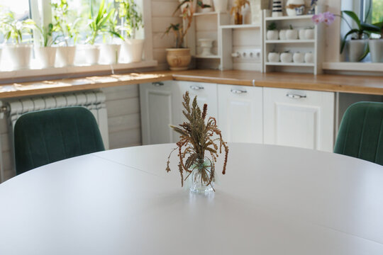 Close view of round white dining table featuring small glass vase with dried brown grasses at center, set inside bright modern farmhouse kitchen with wooden countertop, white cabinetry, potted plants
