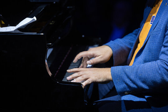Cropped view shows musician hands pressing keys on polished grand piano while folded sheet music rests on open lid, warm stage lighting adding contrast, texture, and live concert atmosphere