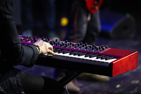 Hand of keyboard player pressing keys on red electric keyboard during performance, with visible knobs and sliders, shallow focus, blurred stage lights, dark clothing, and energetic concert mood