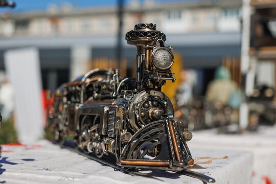 Detailed closeup of metal steam locomotive sculpture showing gears, pipes, coils, perforated headlamp grill, and aged industrial finish on outdoor table in bright sunlight with shallow depth of field