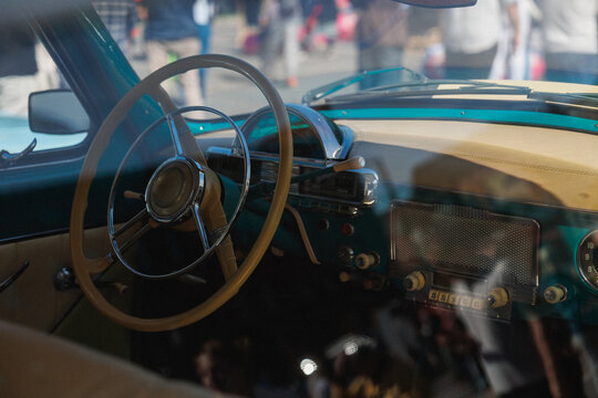 Close-up view through window of vintage turquoise car interior showing large circular steering wheel, chrome horn ring, retro dashboard with round gauges and pushbutton radio controls, cream leather