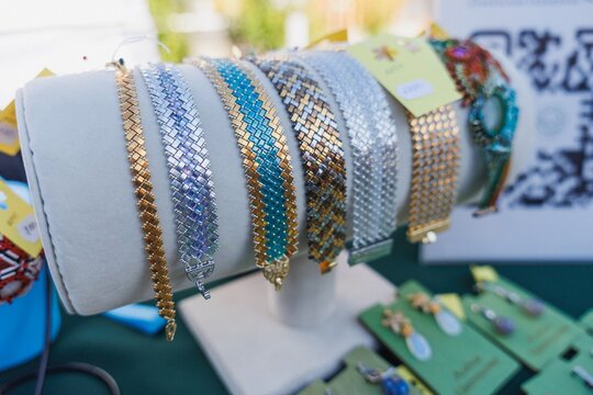 Beaded metal bracelets arranged across padded display stand showing woven link patterns, metallic gold and silver finishes, turquoise and purple accents, visible clasps, textured surfaces, soft