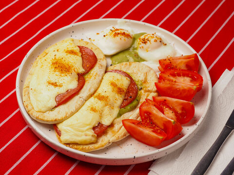 Tasty breakfast plate with Mexican pita bread with German salami and melted cheese, poached fine eggs and sliced fresh tomato. Food served on a red and white stripe table cloth. Huge portion.