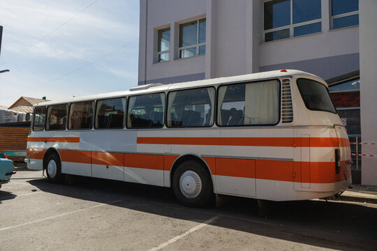 Side view of retro orange and white coach bus parked in lot next to pale modern building with large windows; sunlight casts soft shadows across bus body and asphalt, interior rows of passenger seats