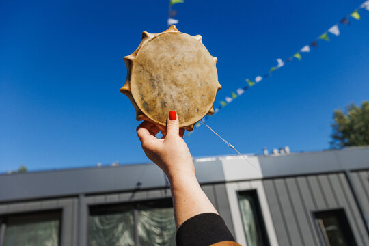 Close-up of hand with red nail polish holding round leather frame drum aloft against deep blue sky, while triangular bunting and modern building facade add festive urban context and bright daylight
