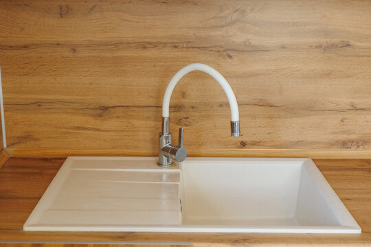 Curved white faucet rises over composite sink and built in drainboard beside chrome base and oak wood backsplash, creating clean modern kitchen utility with soft natural light and closeup detail