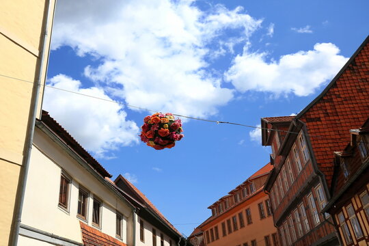 Ein farbenfrohes Blumenarrangement h&auml;ngt an einem Seil &uuml;ber einer historischen Altstadtgasse mit Fachwerkh&auml;usern in Quedlinburg vor einem blauen Sommerhimmel mit wei&szlig;en Wolken.