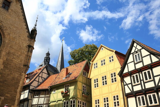 Farbenfrohe Fachwerkfassaden und historische Ziegeld&auml;cher vor der Kulisse von Kircht&uuml;rmen in der Altstadt von Quedlinburg, aufgenommen bei sonnigem Wetter unter blauem Wolkenhimmel.