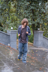 Obraz premium A teenager walks along a wet path under the cloudy sky. Trees surround the area as leaves cover the ground.