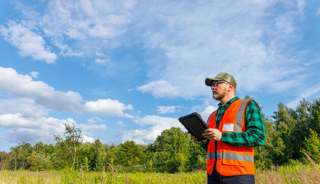 Male ecologist or surveyor wearing an orange reflective vest and cap, holding a digital tablet and looking away during a landscape inspection in a green field. Climatologist.