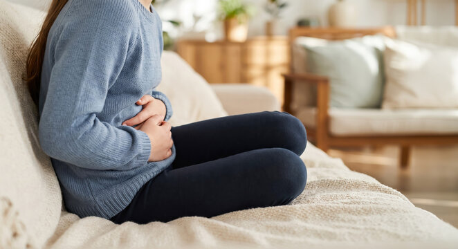 A person on a couch in a living room holds their stomach while looking uncomfortable. Natural light comes through the window. The room has plants and soft furniture