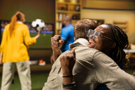 Joyous soccer fans embrace on couch, pleased by positive football match result. Close up of euphoric supporters hugging, celebrating team success during home TV watch party.
