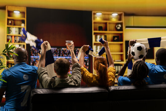 Passionate football fans watching TV, thrilled by positive live soccer match result. Back excited supporters at home raising team colors scarves, doing goal celebration, delighted by victory.
