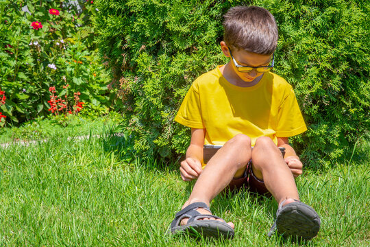 Boy sitting on the lawn in a park with tablet reading electronic book
