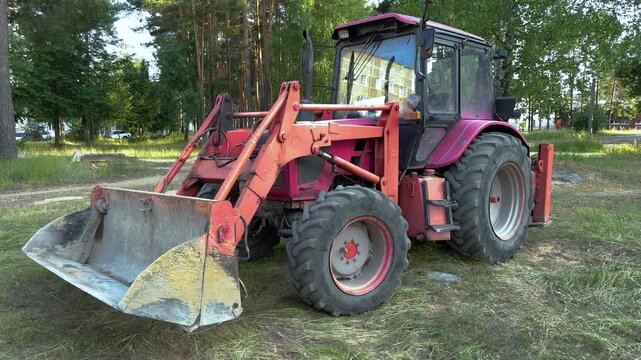 Pink and red tractor with front loader bucket parked on grass near forest. Agricultural machinery, construction equipment, outdoor scene
