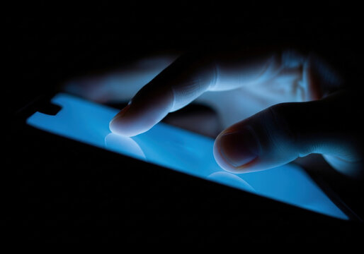 Close up view of person using mobile phone in dark room with fingers touching glowing blue screen representing digital communication and tech.
