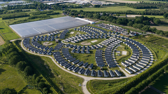 Aerial view of a holiday park with rows of mobile homes arranged in a circular pattern, surrounded by green fields, trees, and a large greenhouse in Venlo, Limburg, Netherlands.