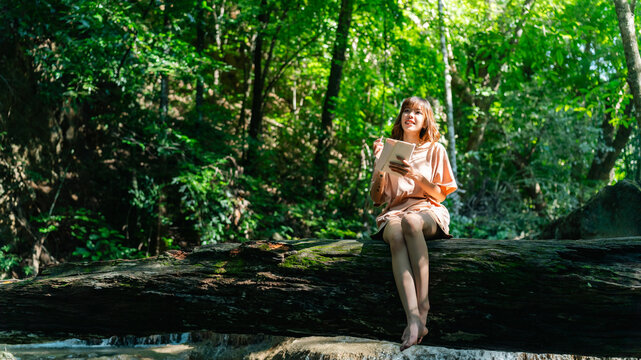 Young Asian woman writing travel journal while relaxing on fallen tree in tropical forest enjoying peaceful nature therapy forest bathing during eco tourism adventure and outdoor wellness escape