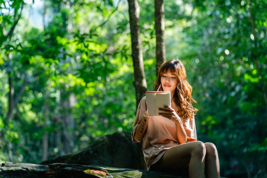 Young Asian woman writing travel journal while relaxing on fallen tree in tropical forest enjoying peaceful nature therapy forest bathing during eco tourism adventure and outdoor wellness escape