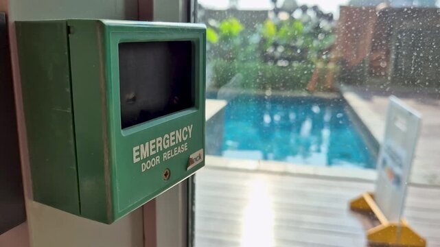Green emergency door release button box on a wall near a blurred outdoor swimming pool.