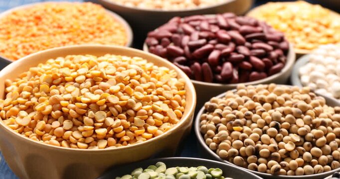Different types of legumes in bowls on blue wooden table, spinning. Closeup view