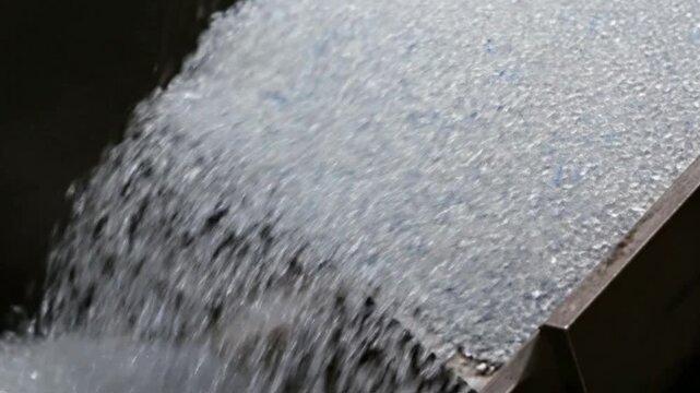 Clear granular material, such as plastic pellets or recycled glass, flowing down an industrial metal chute, illustrating efficient material processing
