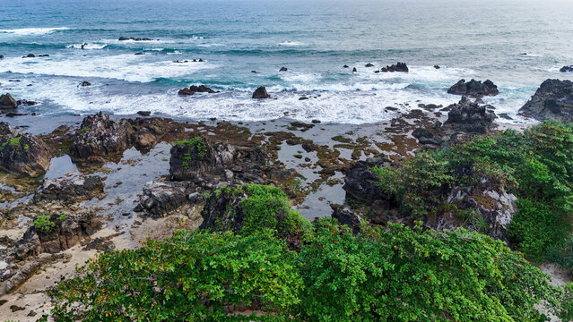 Aerial view of a rocky beach with tide pools, lush green vegetation, and ocean waves crashing against the shore in Indonesia.