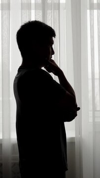 Dramatic silhouette of thoughtful adult man standing near sheer curtains at home. Vertical video of anxiety, uncertainty, loneliness, depression, decision making and emotional pressure concept.