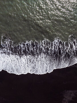 Aerial view of white sea foam crashing onto the dark black sand beach with rhythmic waves and contrast between the dark volcanic sand and the Atlantic Ocean in Iceland.
