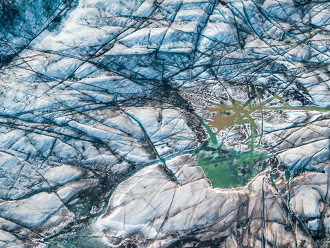 Aerial view of a glacier in Iceland featuring deep crevasses and a vibrant green meltwater pool.