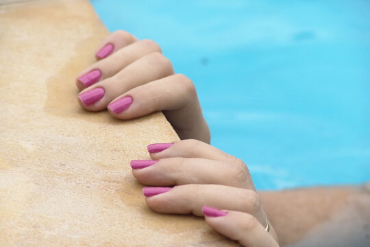 Close-up of a woman's hands with pink painted fingernails holding onto the edge of a swimming pool. - Close-up das m&atilde;os de uma mulher com unhas pintadas de rosa segurando a borda de uma piscina.
