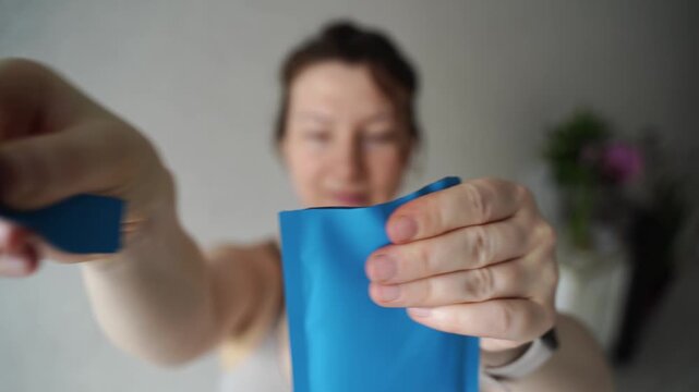 Static Shot Of Woman Tearing Open Blue Sachet And Removing Contents In Indoor Home Setting With Soft Natural Light And Blurred Background.