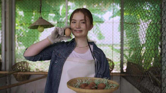 Happy Caucasian woman farmer with an apron standing holding and showing a fresh chicken egg in henhouse. White female teenager is collecting eggs into a basket from a chicken coop on an organic farm. 