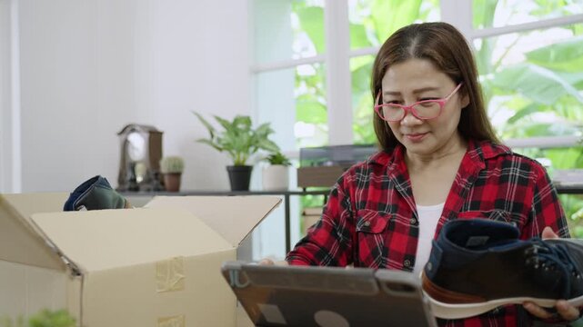 Asian woman entrepreneur, small business owner, with eyeglasses, using tablet to check list of sneakers to delivery from online shopping for customer in home office. E-commerce and e-business concept.