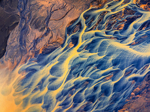 Aerial view of a glacial river delta with intricate blue and yellow sediment patterns flowing through volcanic terrain in Iceland.