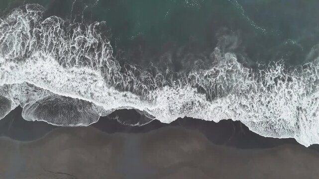 Top down drone view of powerful ocean waves breaking along a black sand coastline, creating striking white foam patterns and generous copy space for text or design.