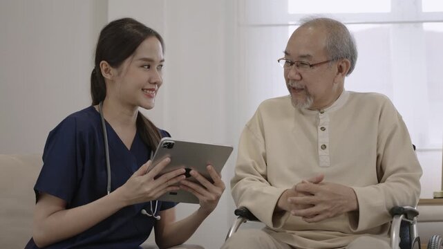 Professional young Asian woman nurse caregiver using a digital tablet to talk and provide advice about healthcare to a senior man patient while sitting in a wheelchair in a special room at hospital. 