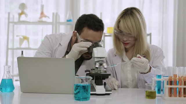 Two happy professional scientists and researchers in lab coats using a microscope and a laptop while conducting chemistry experiments with liquid in test tubes and beakers in the science laboratory. 