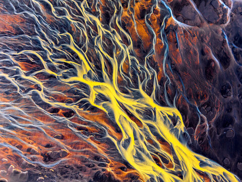 Aerial view of a vibrant braided river system featuring striking yellow and blue glacial sediment patterns flowing through dark volcanic terrain Iceland.