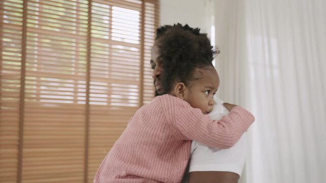 Happy African American father standing, holding, and embracing his cute little daughter with love and bonding in living room at home. A moment of love. A black dad carrying a child girl, affectionate.