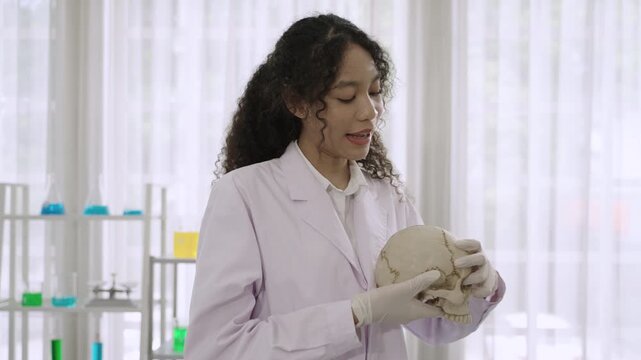 Happy professional woman medical anatomist wearing a white lab coat, standing and holding an anatomical model of the human skull, with a presentation and explanation carefully detailed in laboratory. 