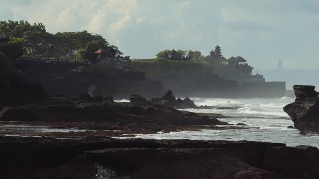 Temple Silhouette Above Jagged Foreground Rocks, Mist Drifting From Surf Over Layered Basalt Ledges Distant Shrine Spire Punctuates Horizon, Cinematic Mood Suited For Pilgrimage, Tanah Lot, Bali