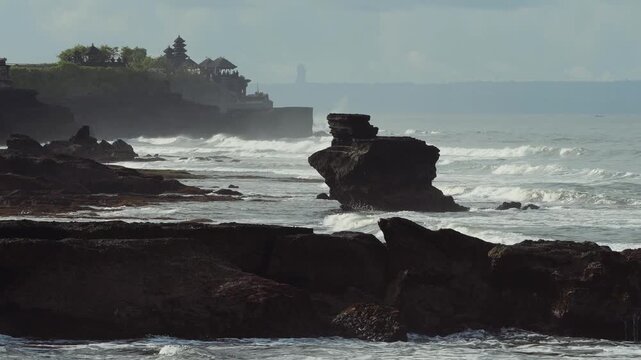 Rock Formation Framed By Surf,Towering Sea Stack With Pounding Waves,Mist Veil And Fierce Spray,Distant Temple Pagoda Tanah Lot On Cliff, Moody Light, Geology And Erosion Focus For Travel Documentary.