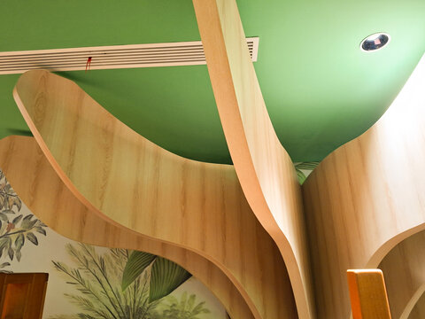 Abstract, wide, curved wooden panels extending from a pillar to form stylized tree branches against a bright green ceiling with ventilation grilles.